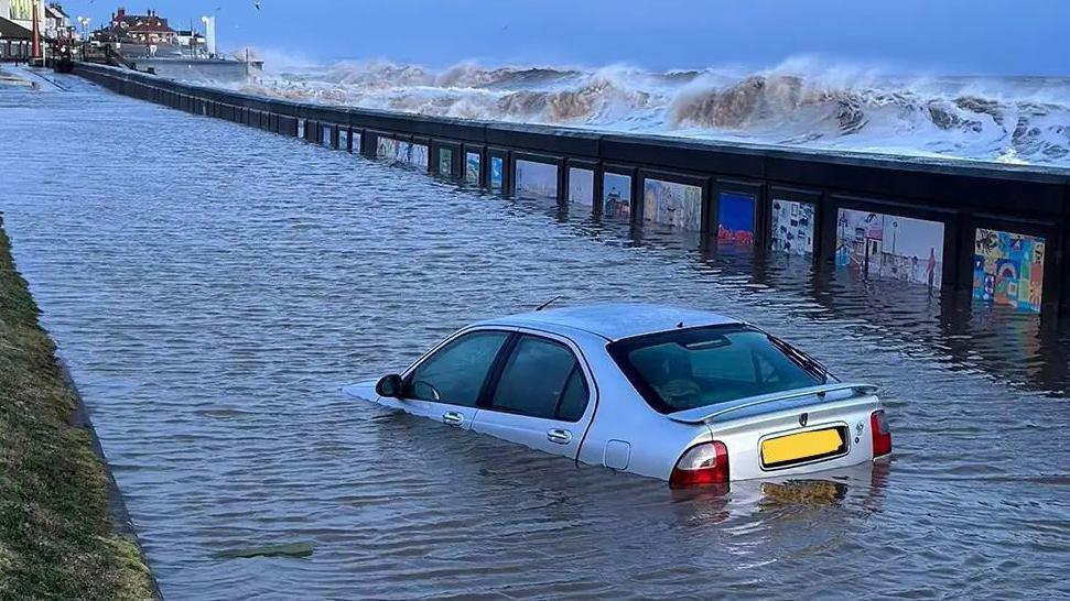 A car is submerged in water along Hornsea seafront. Large waves can be seen on the beach behind the seawall.