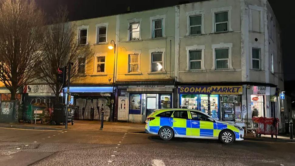 A row of shops at night. A police car is parked at the side of the road. Some of the shops have their shutters up.