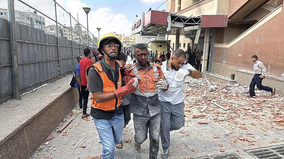 An injured emergency worker is led away by two other emergency workers. There is rubble and wreckage behind them. He is badly hurt.