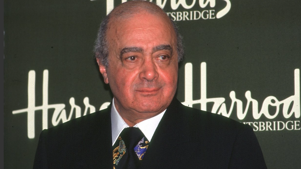 Mohamed Al Fayed wears a dark suit and stands in front of a dark green sign which reads 'Harrods, Kinightsbridge'.