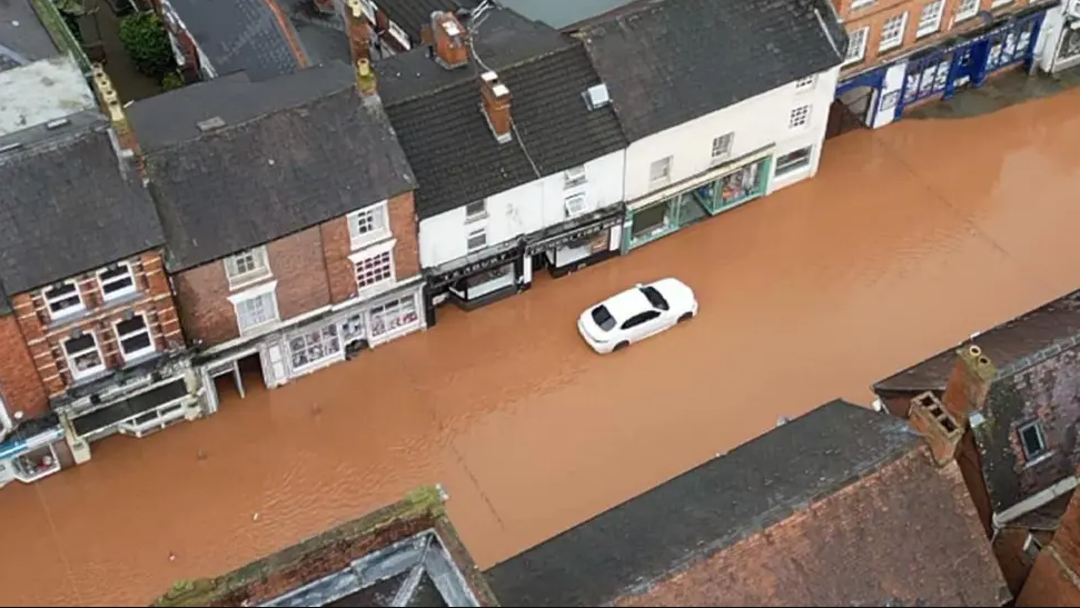 An overhead shot looking down on a street flooded with muddy water. A white car sits in the water. The street is lined with shops and it is clear that the water is very high.