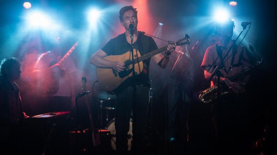 Damian Lewis playing guitar backed by his band