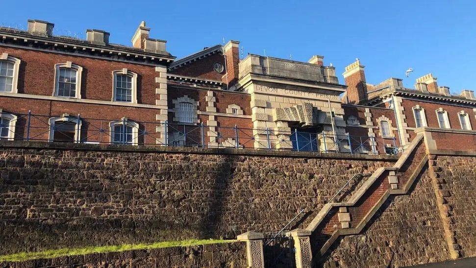 The exterior of HMP Exeter. It is a historic looking building of red brick with ornate granite block work and many chimneys. There is an inscription engraved in the stone above the entrance which reads: DEVON COUNTY PRISON. In the foreground is a stone wall with steps running through it.