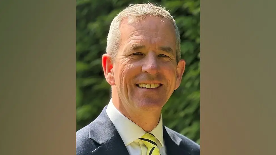 A headshot of a man with short grey hair who is wearing a navy suit and a white shirt with a yellow and navy striped tie.