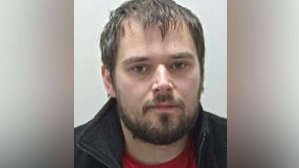 Jordan Blenkinship with dark brown hair, dark brown beard and brown eyes wearing a red t-shirt and black fleece in a police mug shot.