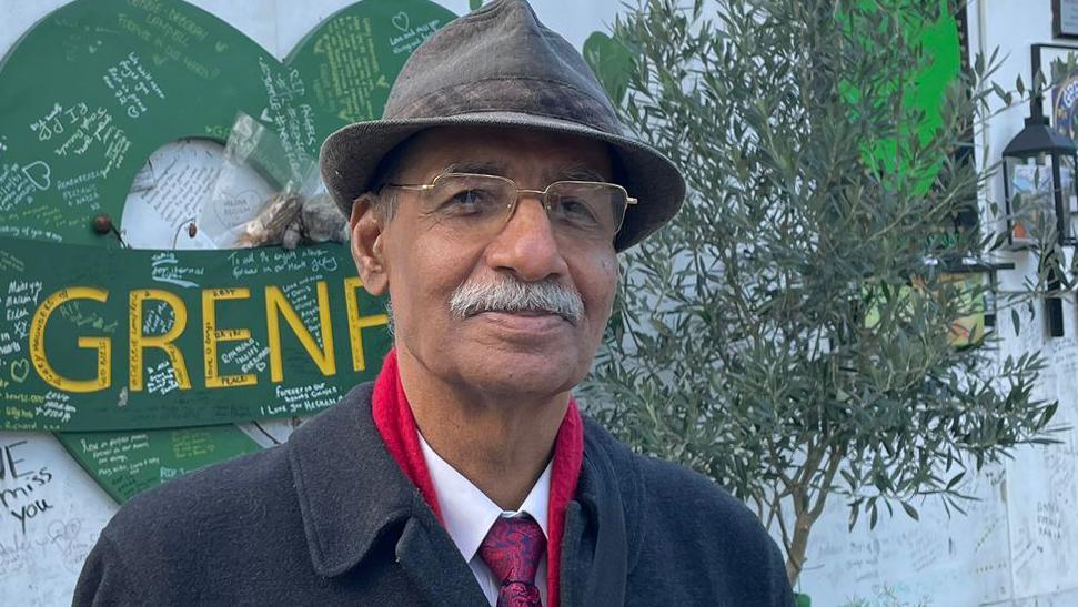 Mushtaq Lasharie is seen wearing a hat, glasses, and a black coat stands in front of a Grenfell memorial wall covered in messages, green hearts, and tribute decorations.