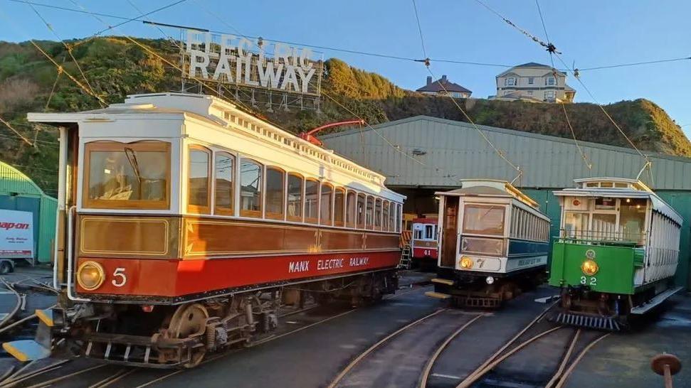 Three Manx Electric Railway cars lined up alongside each other on separate tracks at the depot. The first is red and has the number five, the second is blue and is numbered seven and the third is green and is number 32.