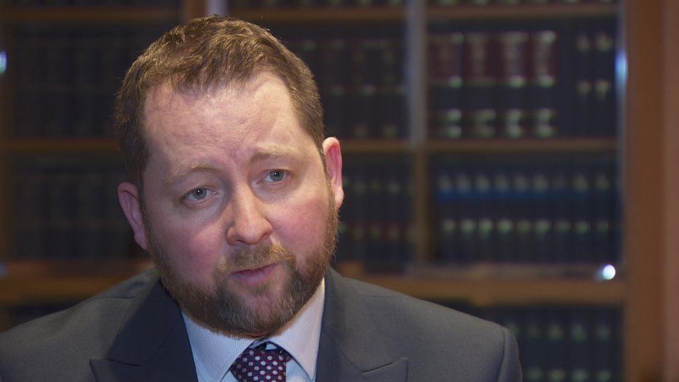 Man in suit and tie with beard in front of library of books 