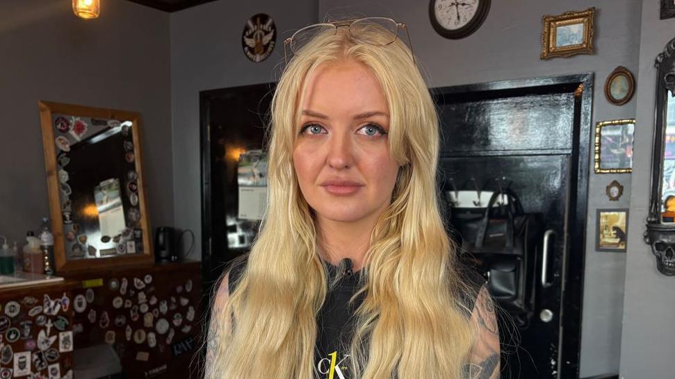 A woman, with long blond hair, standing in a room with grey walls. There is a black door behind her and a number of frames and a clock on the wall. She is wearing a black top with no sleeves. She has tattoos on her arms and glasses sitting atop her head.