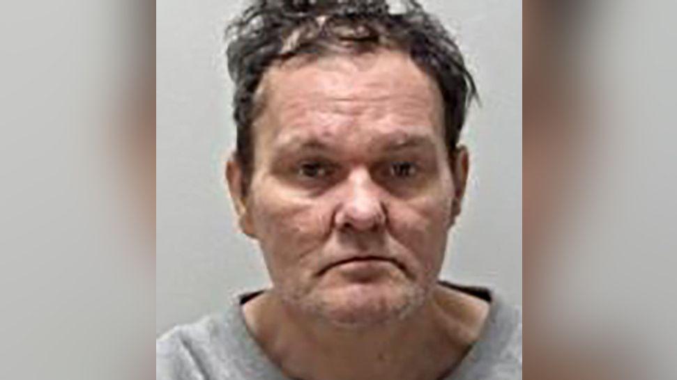 Police mugshot of Adam Saunders, who has brown unruly hair. He is photographed wearing a grey top.