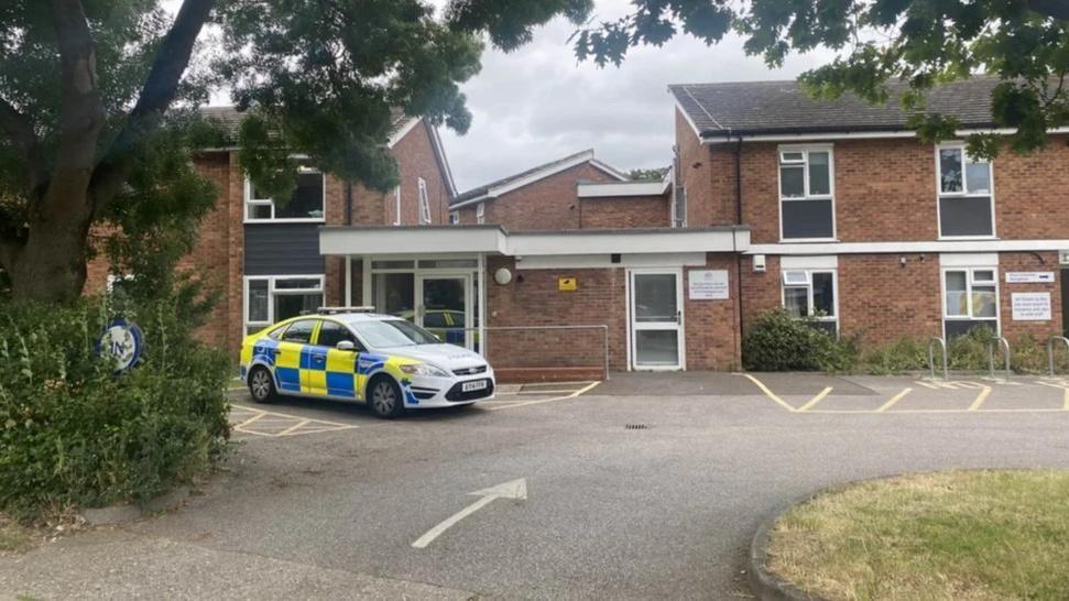 Police car parked outside a red-brick, double-storey housing complex. The is a car park to the front of the building and a green patch of grass. 