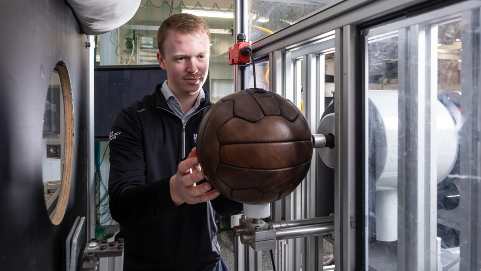 Phillips holding a brown leather ball and placing it into a metal contraption. He has blonde hair and is wearing a black jumper. 