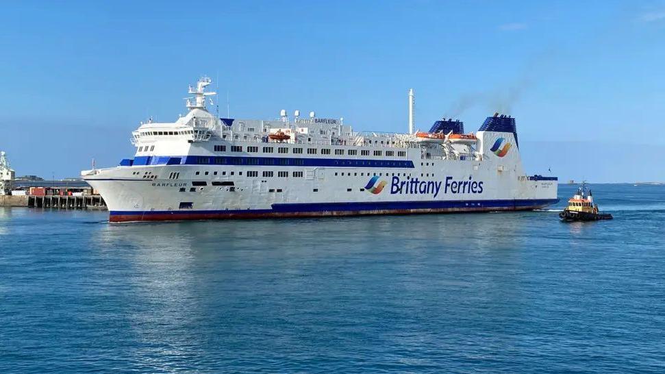 A large Brittany Ferries vessel with a tug to its side and a harbour wall on the other side.