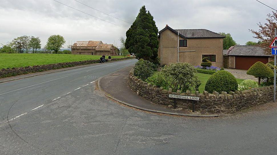 Junction of Ribchester Road and Hothersall Lane in Ribchester. It shows Hothersall Lane road sign and low stone wall in the garden of a house with a large tree to the left and people walking on the pavement of Ribchester Road with a house having a new roof being built in the background.