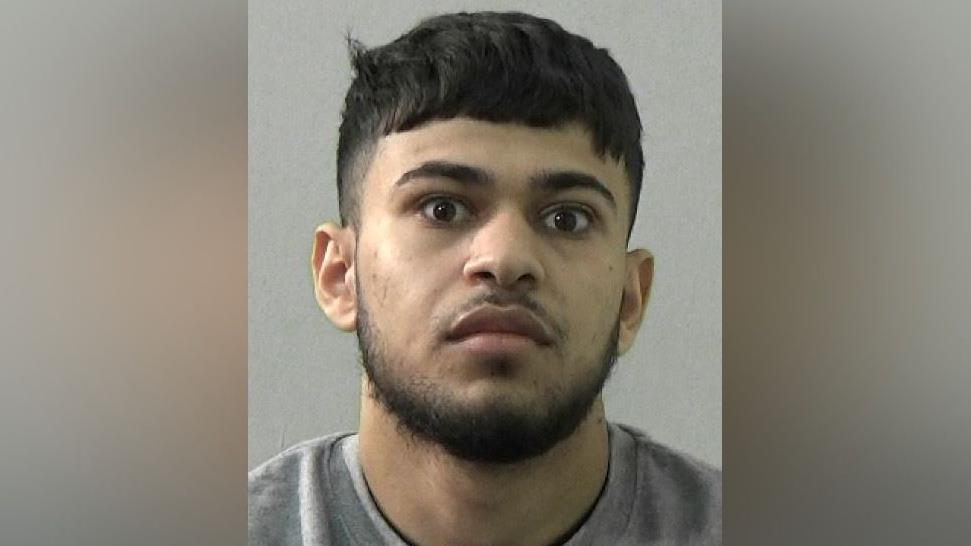 Mugshot of a man with a black beard and hair and wearing a grey sweatshirt