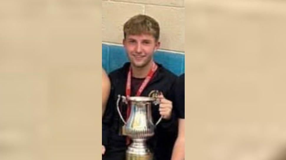 Cain Pears is smiling at the camera as he holds a silver trophy cup. He has short blonde hair and is wearing a black sweatshirt.