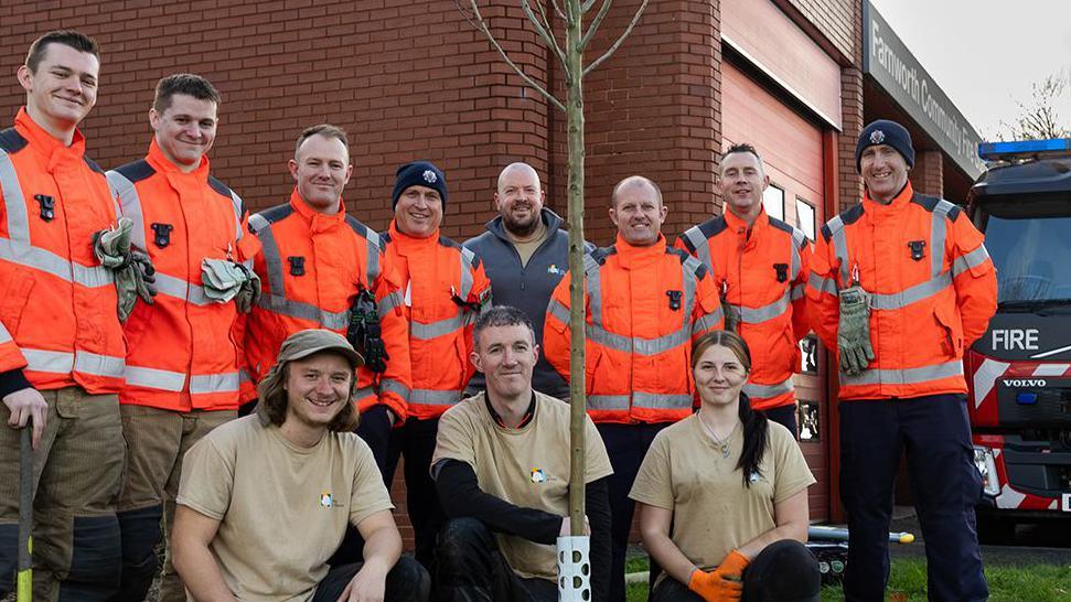 City of Trees volunteers kneeling down in front of a tree planted at with Farnworth fire station with firefighters standing behind them and a fire engine in the background. 