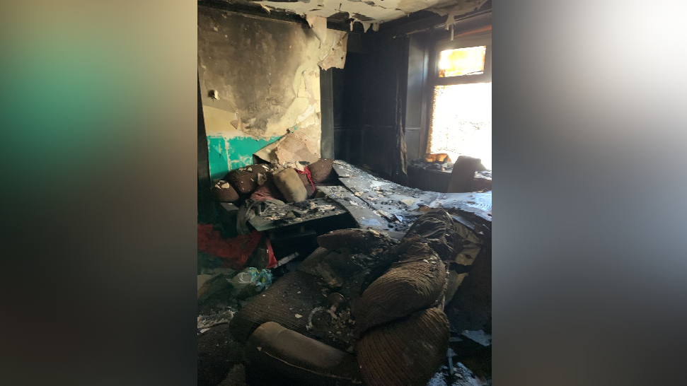 The inside of the same terraced house in another room, with new debris including a burnt and dirt covered sofa and peeling burnt wallpaper 