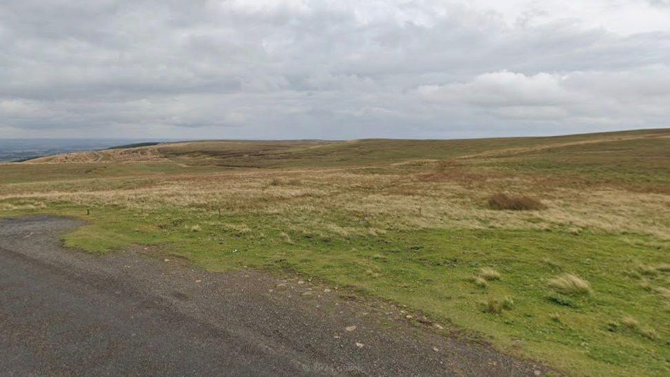 The photo shows the edge of a road in the foreground and rolling moorland stretching into the distance under grey, cloudy skies