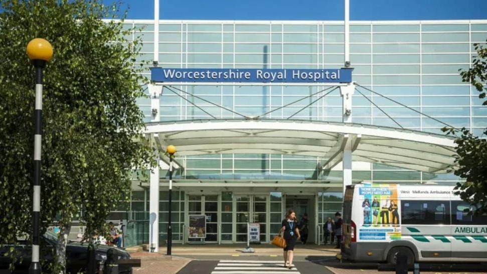 The main entrance of the Worcestershire Royal Hospital building, including a sign at the top and an ambulance on the right.