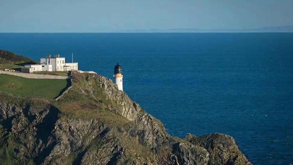 A section of the North East coast of the Isle of Man, you can see a light house on a cliff and the blue sea in the background.