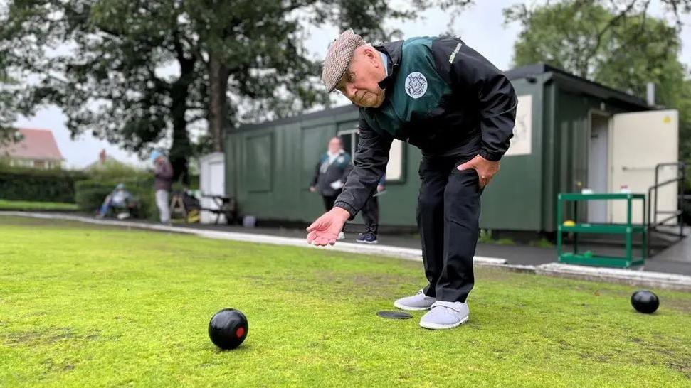 A man playing bowls on a bowling green in Leeds. He is dressed in a black jacket and trousers and is wearing a flat cap.