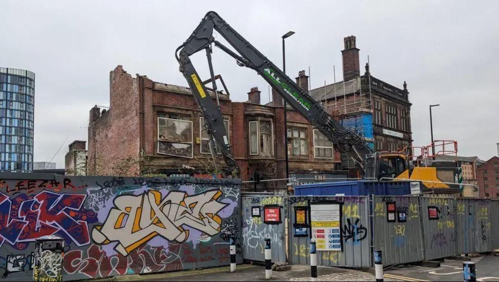 A digger disassembles a large red brick building behind metal screens. The building already has part of its roof and windows missing.