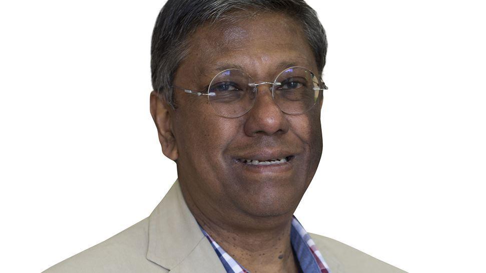 A man with dark hair and wire framed glasses with a blue shirt and pale jacket against a plain white background