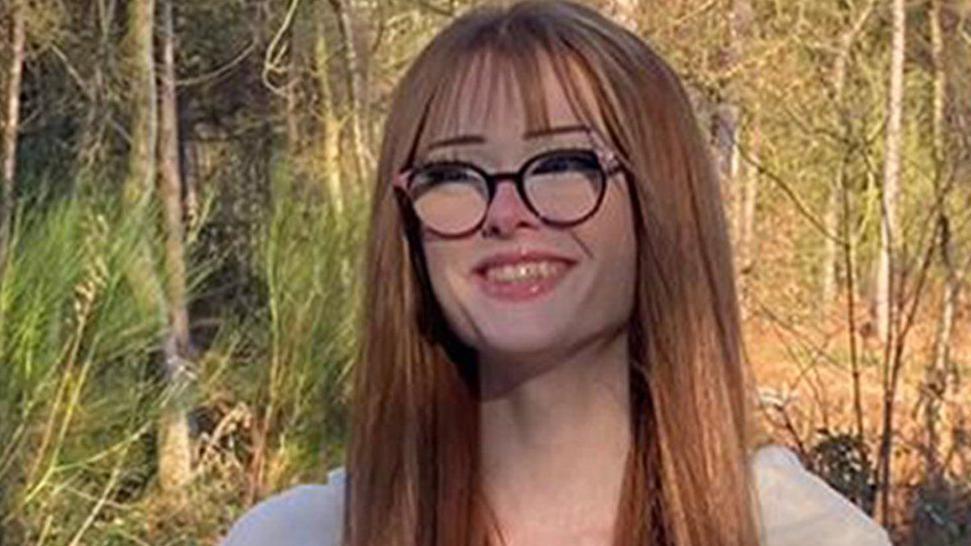 Brianna Ghey, with long ginger hair and wearing wide rimmed black glasses, smiles at the camera against a woodland backdrop