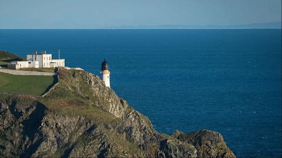 A white lighthouse on a rocky coastline looks out over a dark blue body of water topped with a blue sky. the outline of a neighbouring landmass can be seen in the distance.