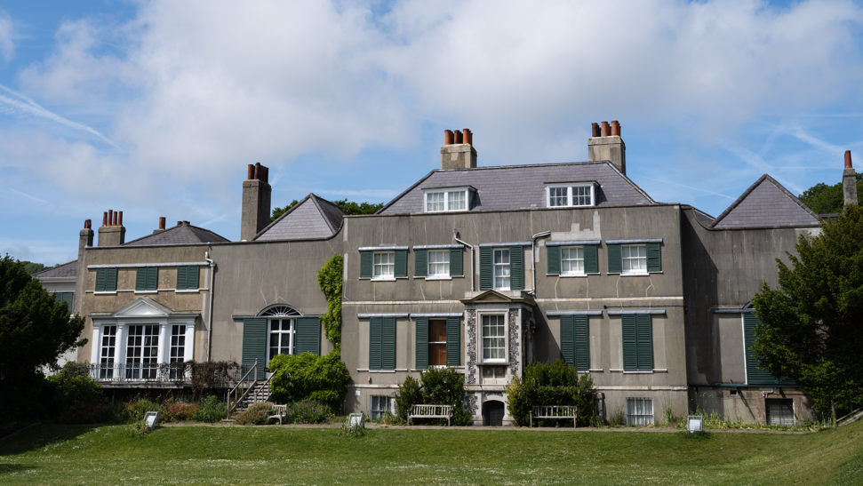 Preston Manor on the outskirts of Brighton. The three-storey manor has white windows and green window covers, alongside a number of visible chimneys. 