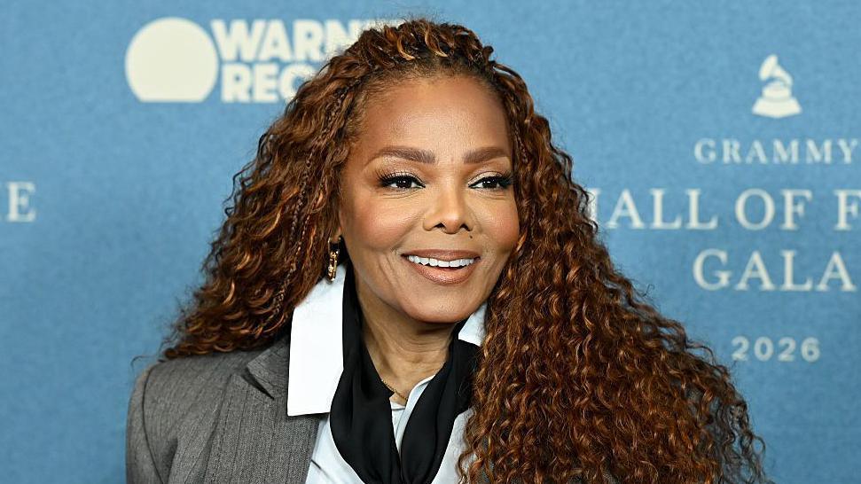 Smiling Janet Jackson stood at a press event against a blue backdrop