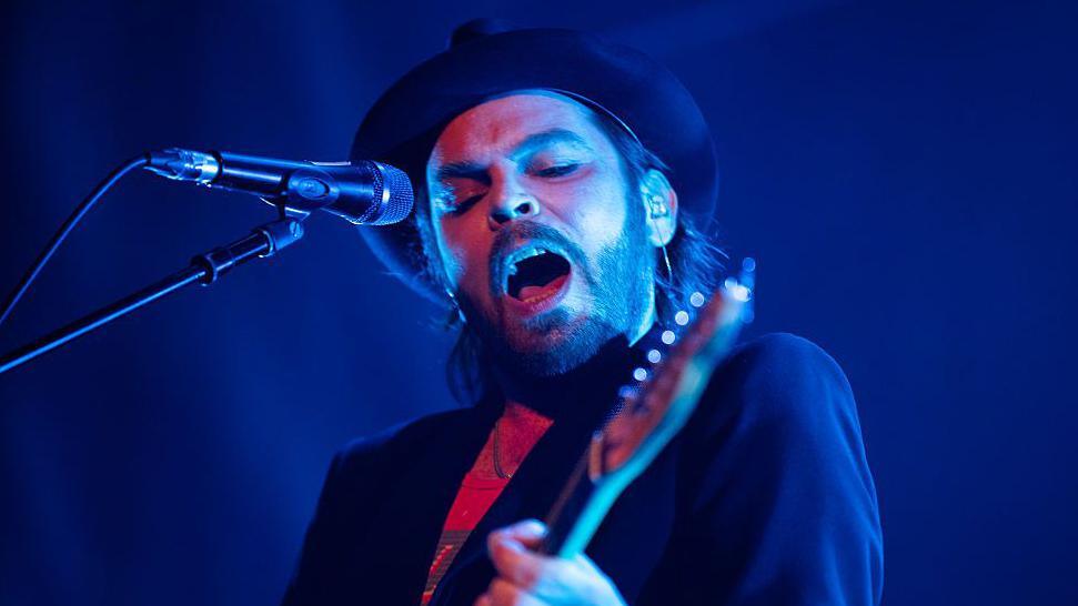 Supergrass frontman Gaz Coombes is wearing a hat and singing into a microphone while playing a guitar on a stage. He is wearing a dark suit jacket.