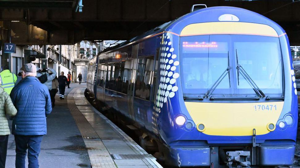 Front view of a train at a station as people walk on platform to the left