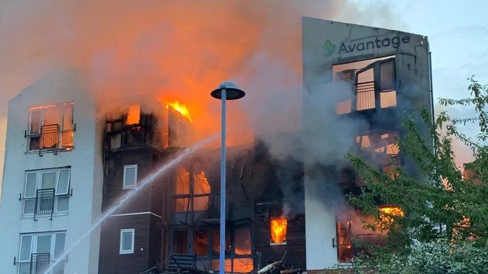 A building which is significantly on fire can be seen with a jet of water targeting an area in the centre while flames are visible on either side