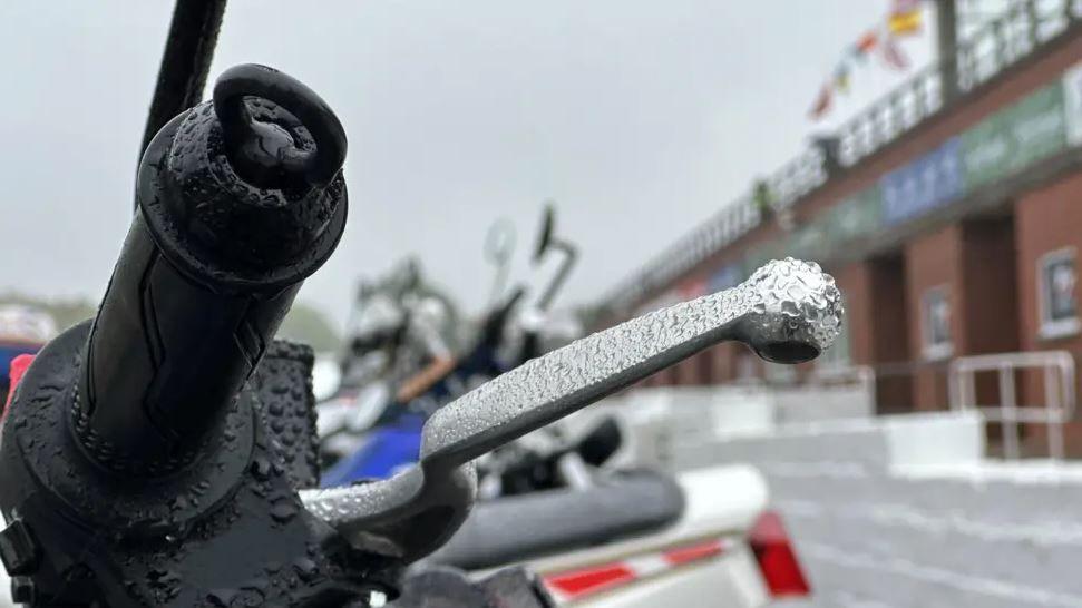 The handlebar and brake lever of a motorbike with raindrops on them. There is a row of other bikes next to it in the background.