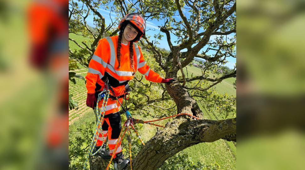 A young woman in orange high-vis clothing stands in the boughs of a tree. She has a harness and ropes and is wearing an orange hard hat. She is smiling at the camera.