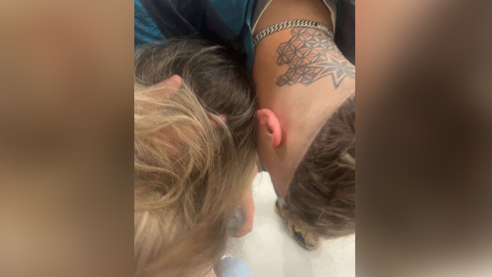 Libby's boyfriend comforting her whilst she is in A&E. Neither of their faces can be seen as both are looking down at the floor. Libby's boyfriend is holding onto her head and trying to give her water.