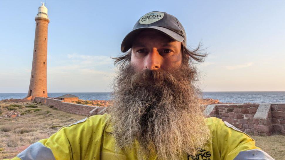 Sandy Duthie, man with large greying beard, yellow Parks Victoria jacket, with a lighthouse in the background, and sea, and blue skies.