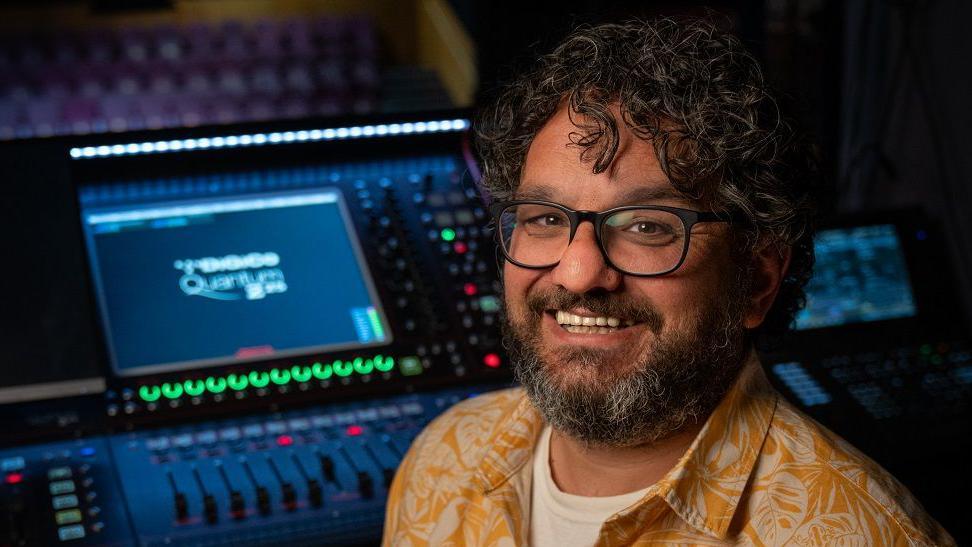 A man with dark curly hair and glasses smiling into the camera in front of a sound deck with lots of switches and buttons. He is wearing a bright yellow, floral shirt.