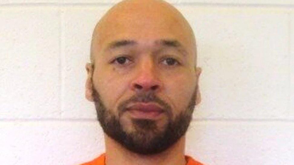 A mug shot of Tremane Wood wearing an orange prison shirt as he stands against a white wall