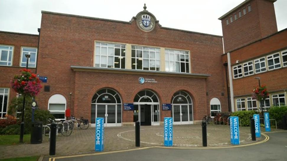 The University of Worcester sign is on the front of a red-brick building with arched entrances and several bike stands in front of the building. A crest is visible at the top of the building.