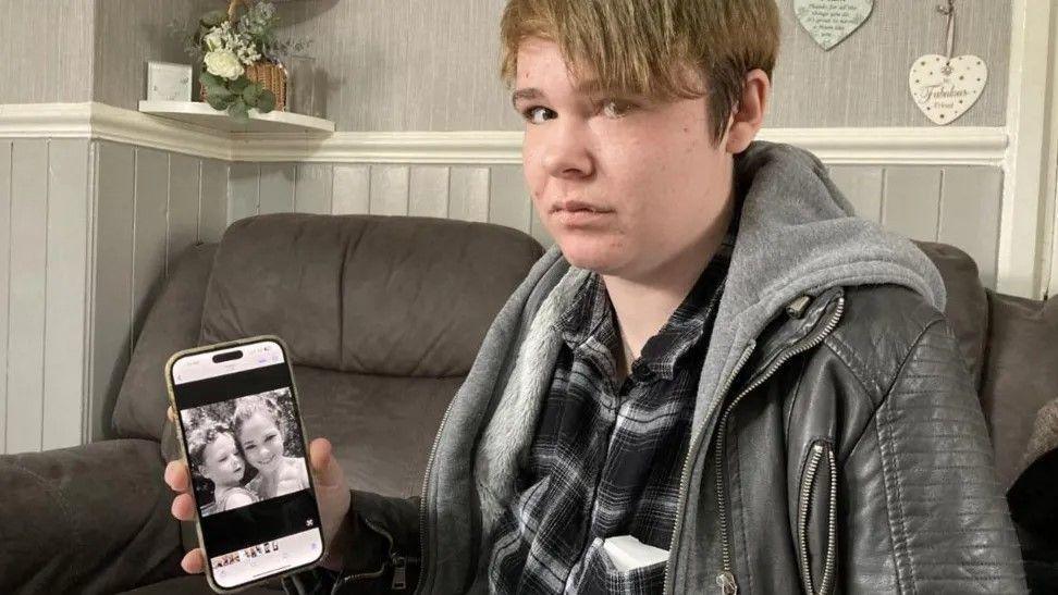 A woman in a leather jacket and checked shirt holds up a phone, with a picture of a woman and a baby boy on it