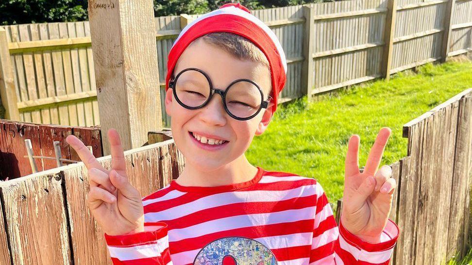 A boy wearing a red and white striped long-sleeved t-shirt and red and white striped hat with round black-rimmed glasses is smiling and holding hands up in peace sign. 