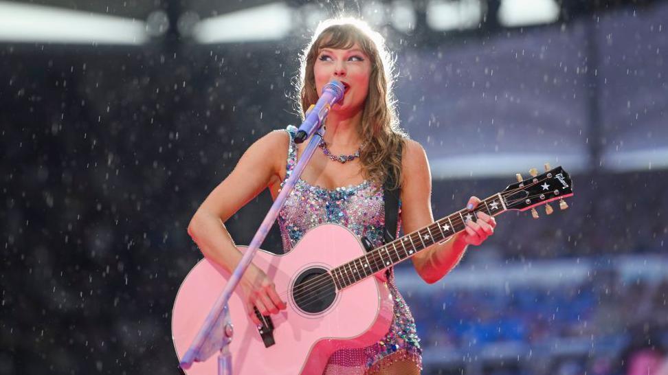 Taylor swift performs in the rain in a sparkly bodysuit whilst playing a pink guitar 