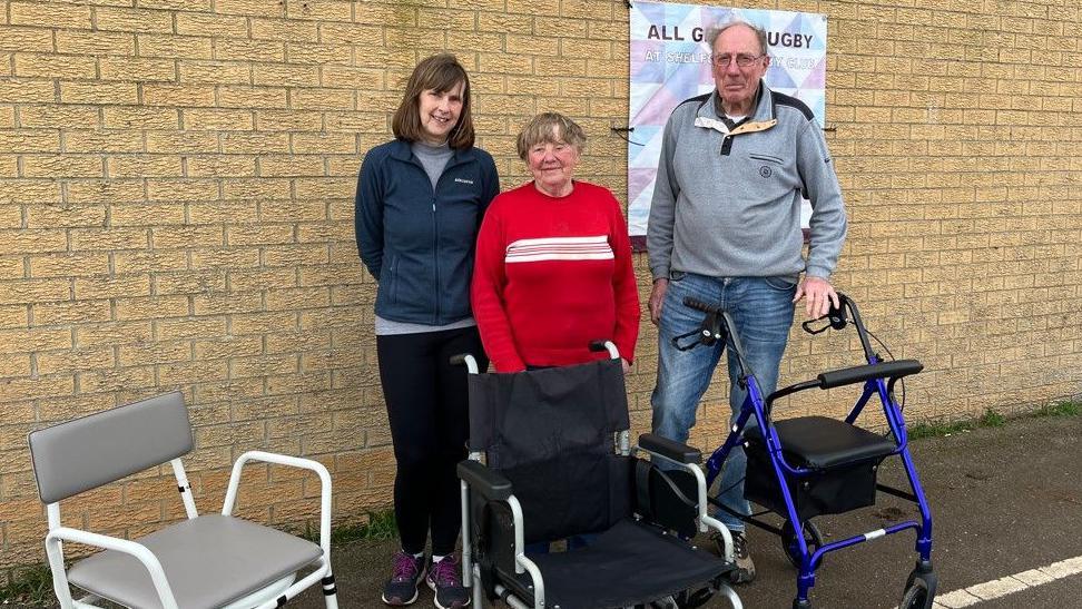 Jo Hutchings, Gaynor Farrant and David Farrant are standing against a brick wall at a rugby club. In front of them they have mobility aids - a commode, a wheelchair and a walking aid.