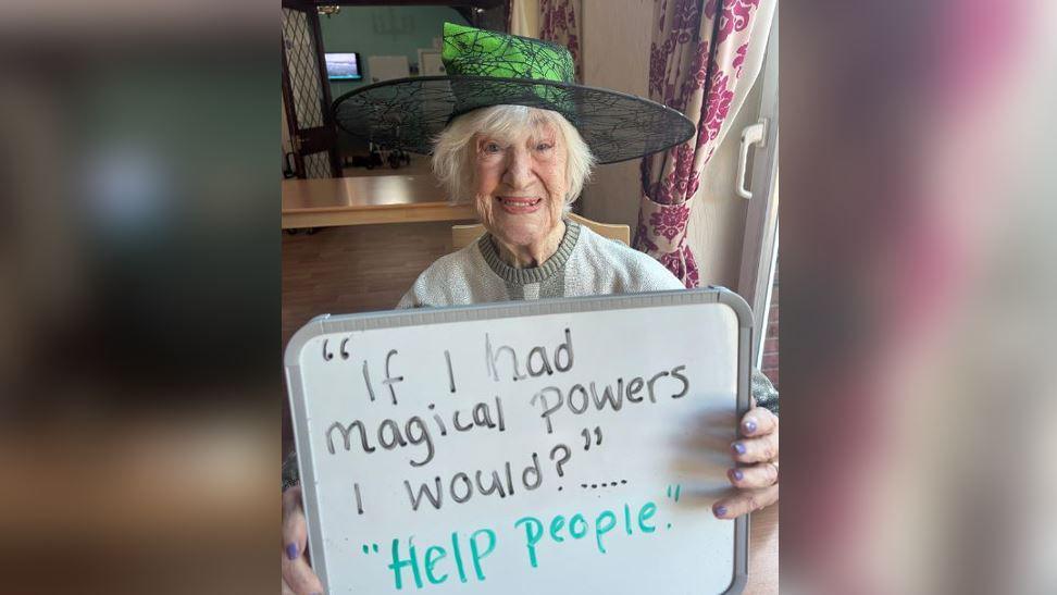 An elderly woman sitting in a chair, smiling, wearing a witches hat and holding up a sign that says "if I had magical powers I would?" with the answer "help people".