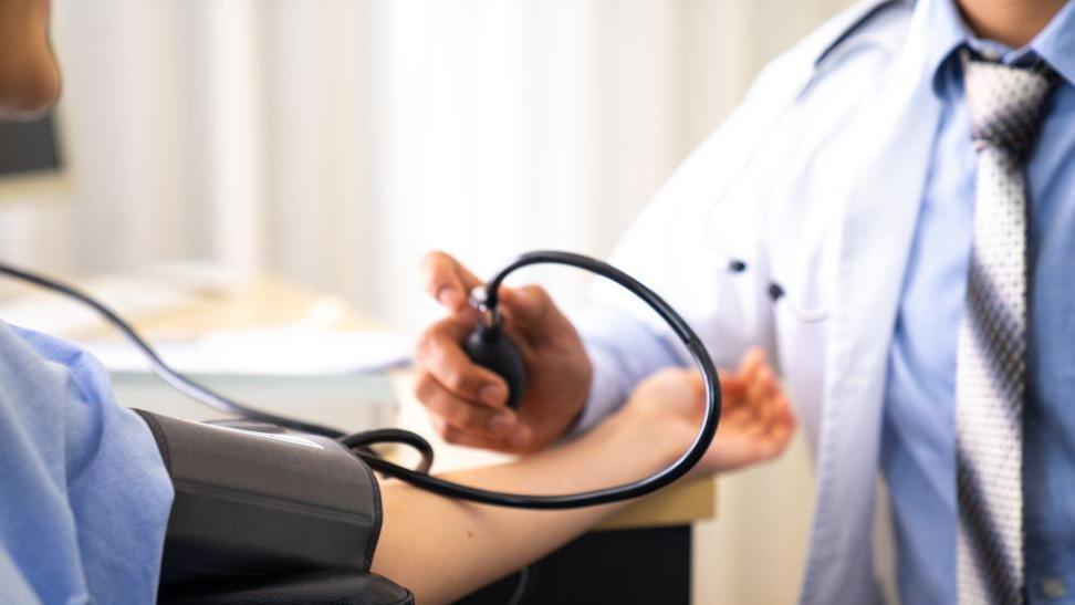 Doctor in white coat, with blue shirt and grey tie takes blood pressure from a patient wearing a blue top.
