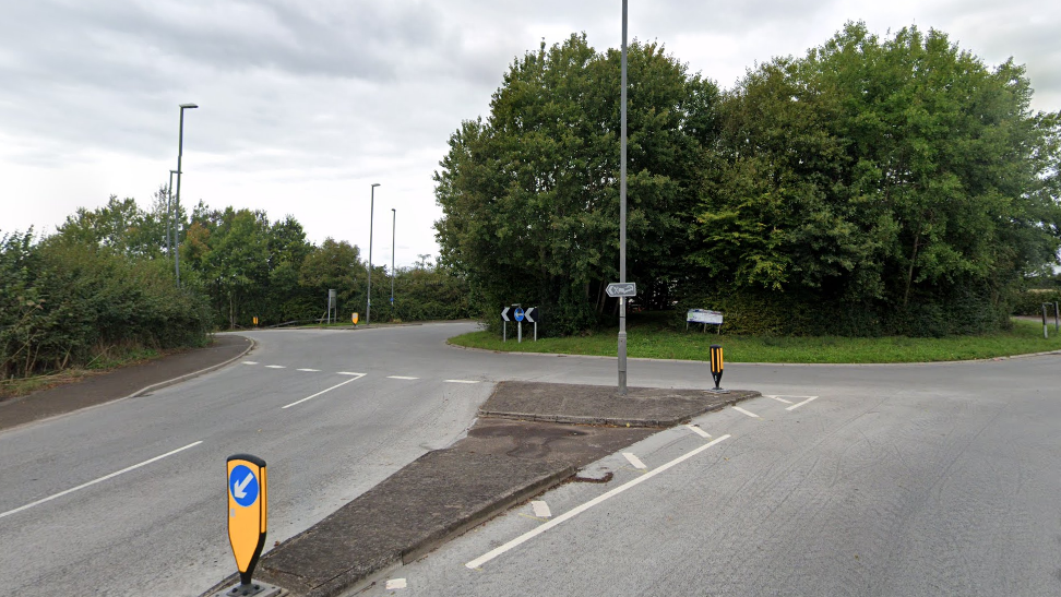A road leading to a roundabout with two green trees in the middle. The sky is grey.