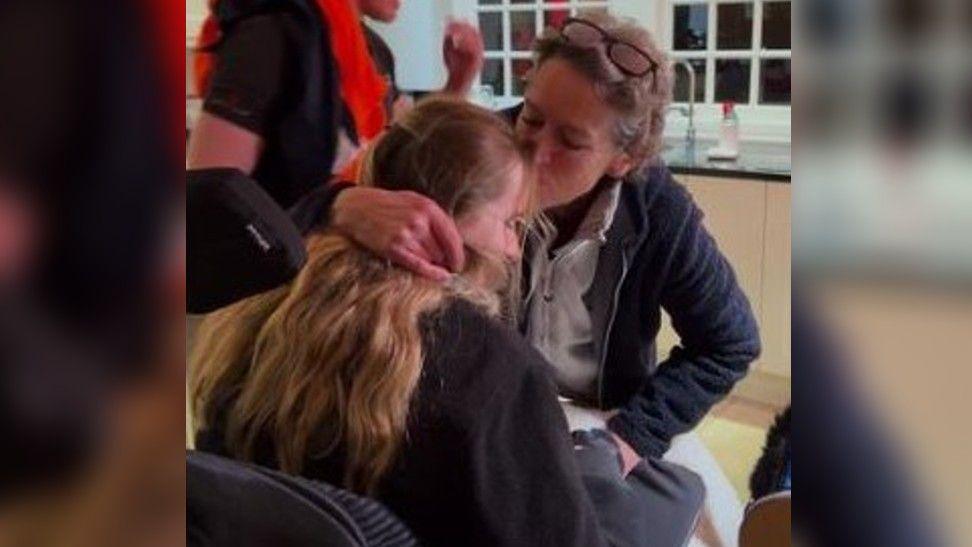 A woman is leaning forward to kiss her daughter on the forehead. The daughter is a wheelchair user. Someone else could be seen behind them.